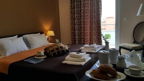 A hotel room with a bed, a bedside table with a lamp, a window with curtains, a chair, and breakfast items on the table. - Yakinthos Hotel (Katerini)