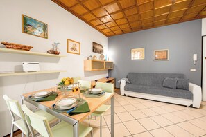 A dining area with a wooden table set for four, a grey sofa, and a kitchen area with a white fridge and shelves. - Il Sogno Di Silvana (Sanremo)