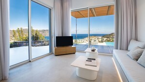 A modern living room with a large TV, a coffee table, and a sofa. It features large windows with a view of the sea and hills. - Istia Luxury Villas (Karpathos)
