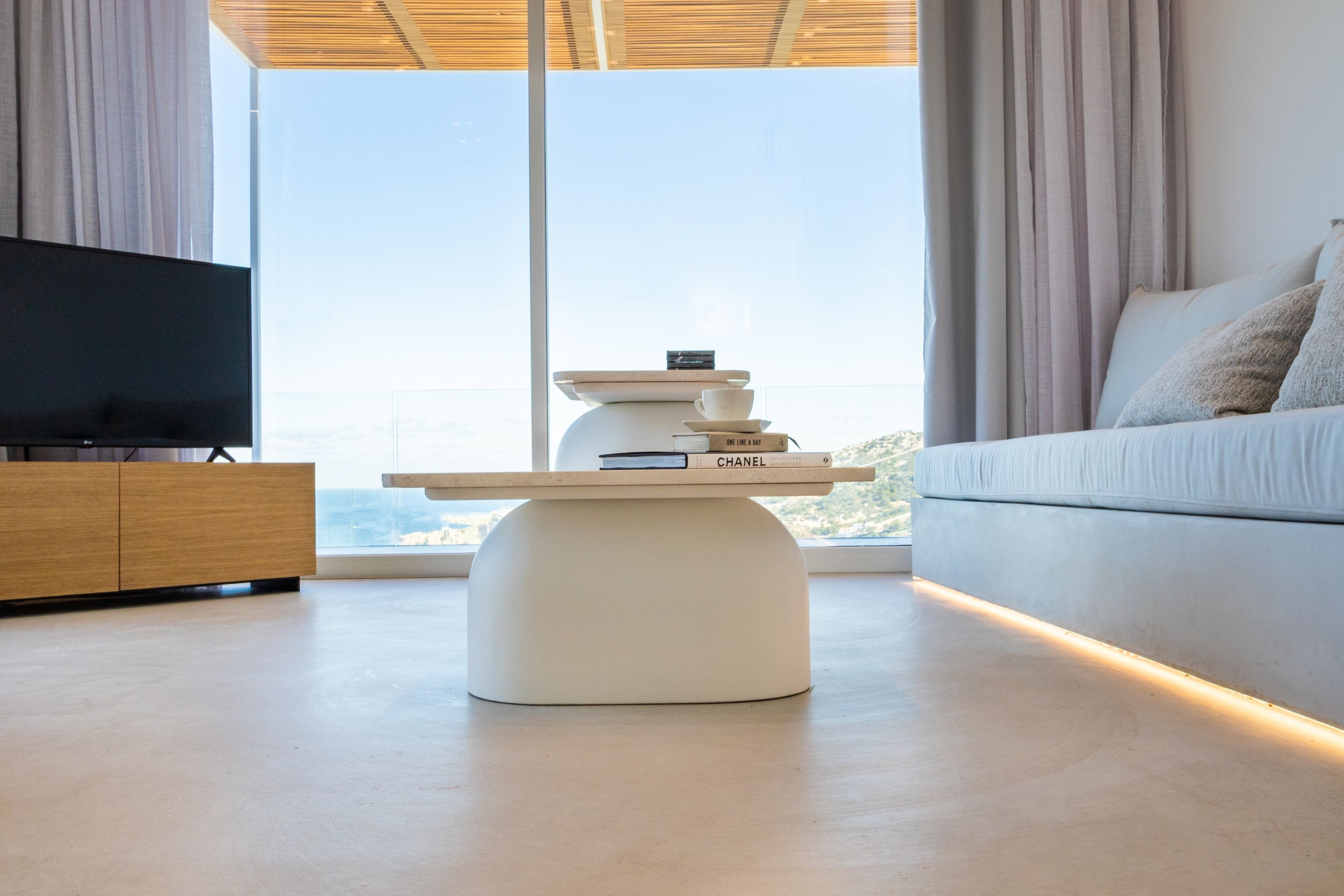 A modern living room with a flat-screen TV, a white coffee table with books, and a sofa with cushions.
