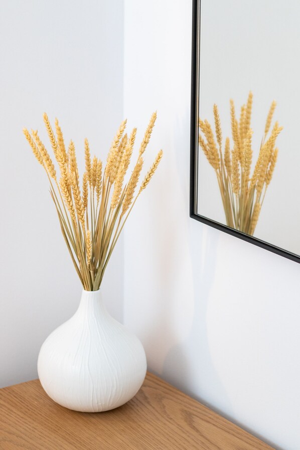 A white vase with wheat stalks on a wooden surface next to a framed picture of wheat on a white wall.