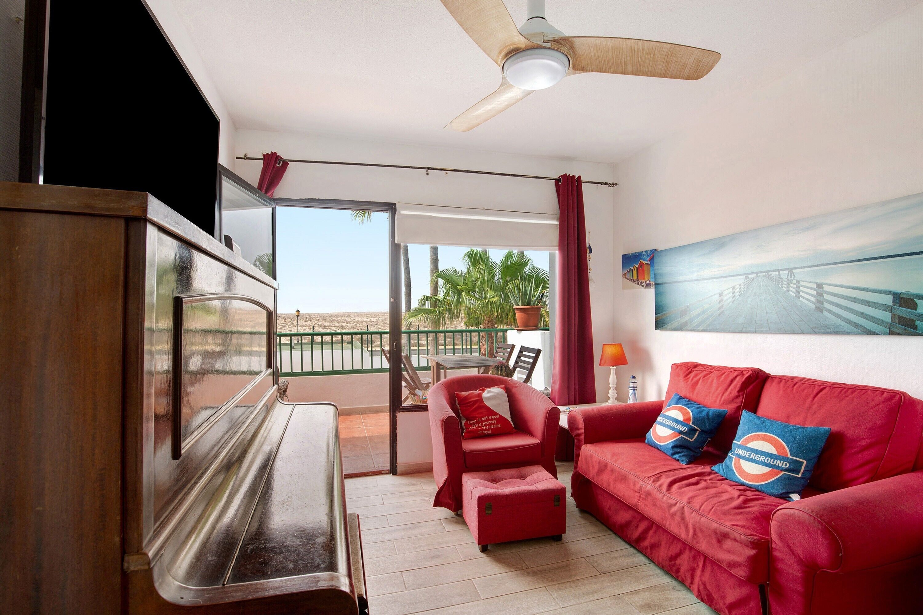 Ein Zimmer mit einem Klavier, einem roten Sofa mit 'Underground'-Kissen, einem Deckenventilator und einem Balkon mit Blick auf Palmen.