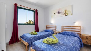 Ein Zimmer mit zwei Betten, einem Fenster mit roten Vorhängen, einer Wandlampe und einem Blumenbild.