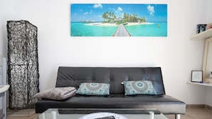 Ein Wohnzimmer mit einer schwarzen Ledersofa, gemusterten Kissen, einer Fernbedienung auf einem Couchtisch und einer Darstellung einer tropischen Strandlandschaft an der Wand.