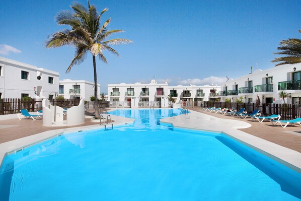 A swimming pool with lounge chairs, palm trees, and white buildings with balconies.