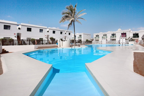 Une piscine bordée d’un palmier, des bâtiments blancs et un ciel d’un bleu limpide.
