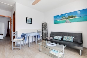 Un salon avec une table basse en verre, un canapé agrémenté de coussins à motifs, un coin repas avec une table et des chaises, et un tableau représentant une scène de plage.
