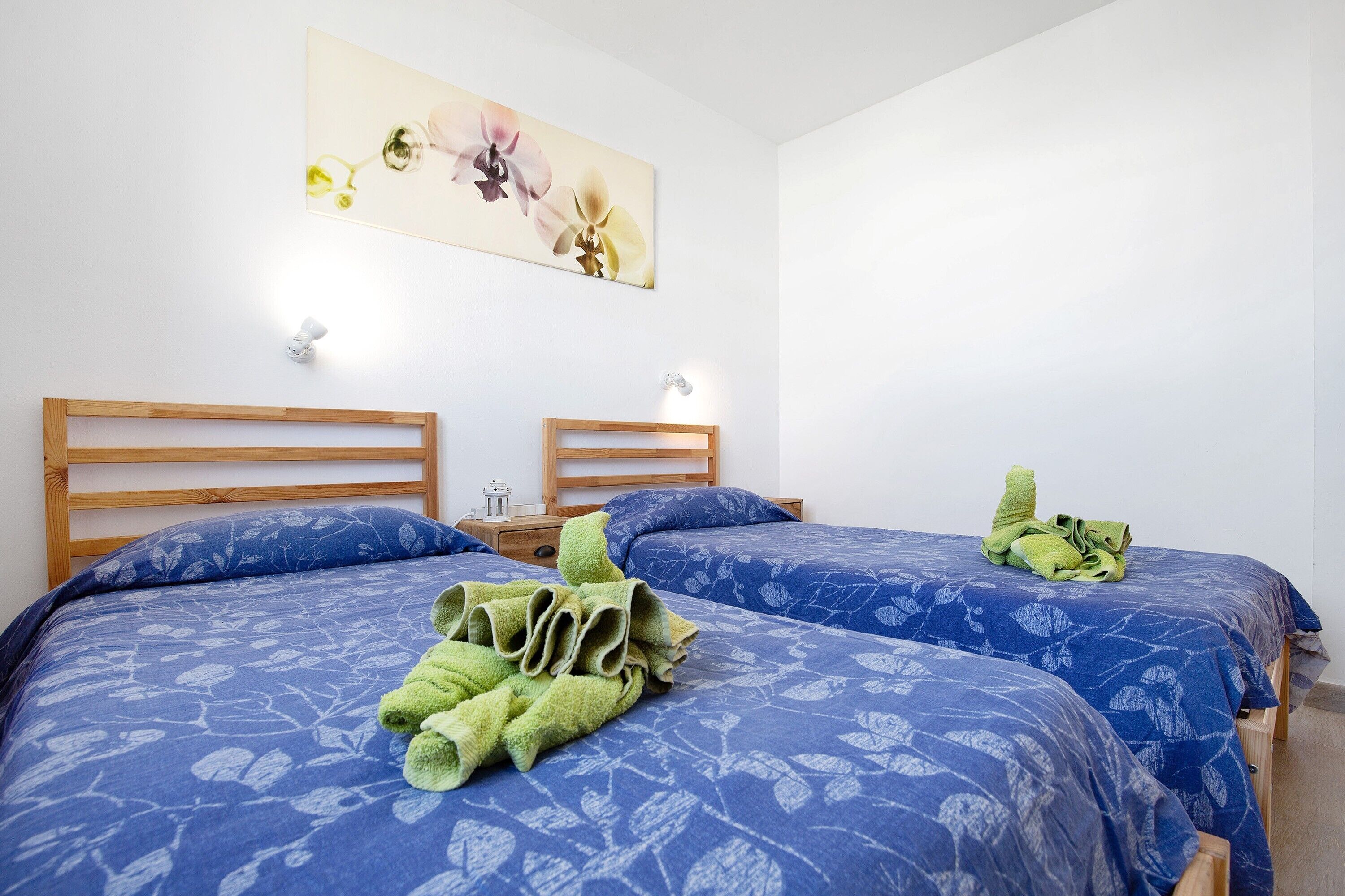 Habitación de hotel con dos camas, cada una con colcha azul estampada con flores, cabecero de madera y mesita de noche con lámpara.