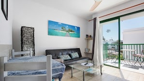 Ein Wohnzimmer mit einer Couch, einem Glastisch, einem Etagenbett, einem Gemälde mit tropischer Szenerie und einem Balkon mit Gartenmöbeln.