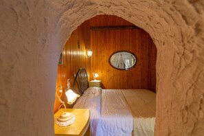 A cozy room with a wooden ceiling, a round mirror, a bed, a nightstand, and a lamp.