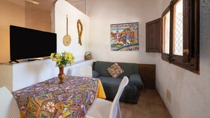 A cozy room with a sofa, a table with a patterned tablecloth, a TV, a wall with a decorative painting, and a window with a lattice design. - Holiday Home 'Fattoria Spezia - Limone' with Shared Pool, Shared Garden and Wi-Fi (Buseto Palizzolo)