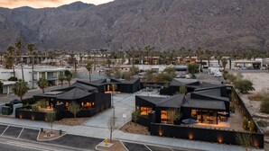 Een modern hotel bestaande uit meerdere gebouwen met zwarte daken, gerangschikt rond een binnentuin met palmbomen en een berg op de achtergrond.