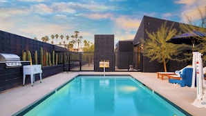 Una piscina con barbacoa y sombrilla, rodeada de palmeras y arquitectura moderna.