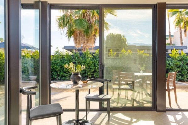 A modern room with a glass door leading to a patio. Inside, there is a round table with two chairs, a vase with flowers, and a small table with a plant.