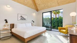 Ein modernes Schlafzimmer mit Holzboden, einem großen Bett, einem Nachttisch mit Lampe, einem gelben Sessel und Blick auf Grün durch eine Schiebetür.