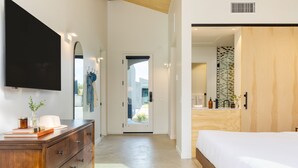 Un dormitorio moderno con una cama grande, un aparador de madera, un televisor de pantalla plana y un baño visible a través de una puerta abierta.