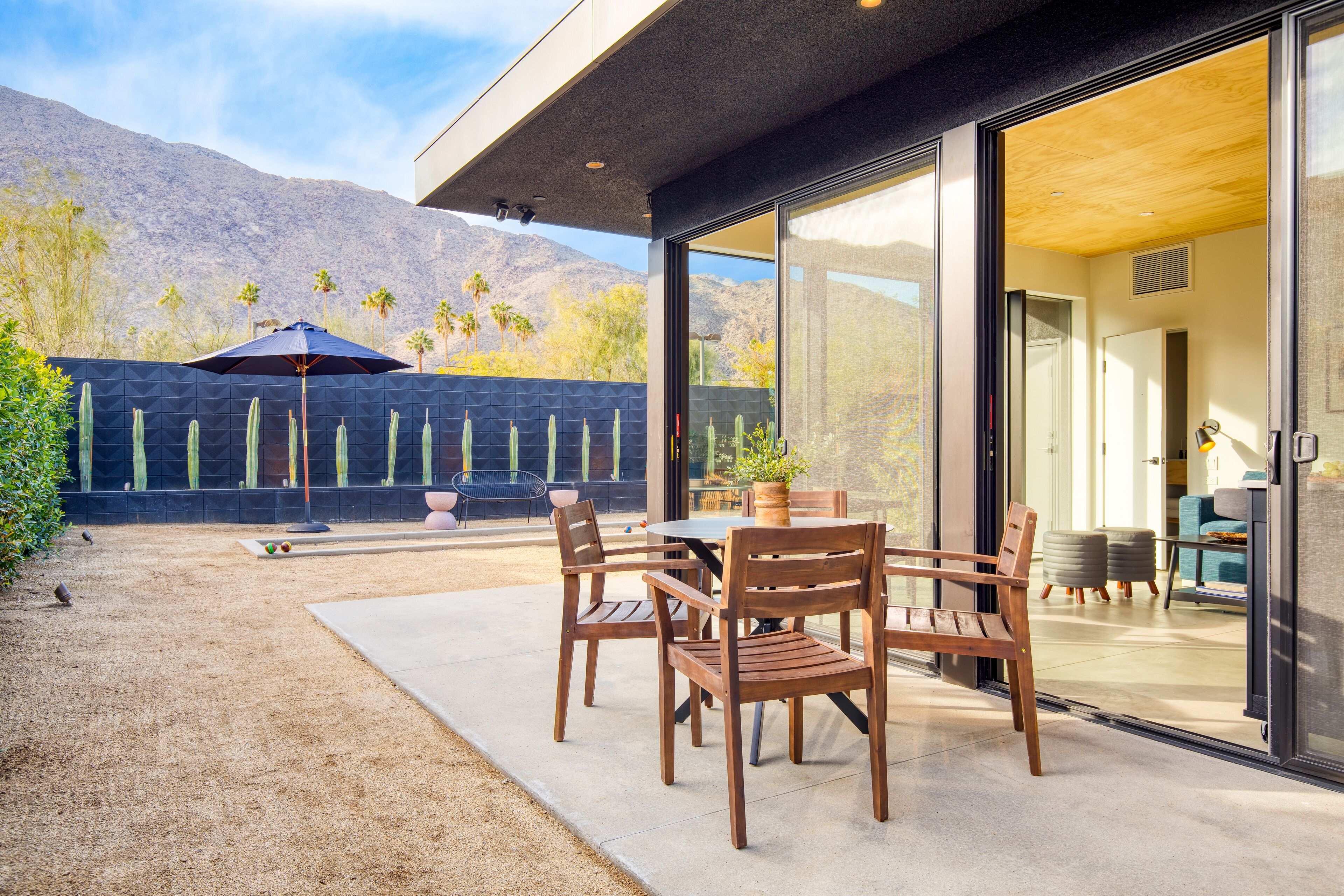 An outdoor patio with wooden furniture, a table, and chairs, a potted plant, and a cactus garden.