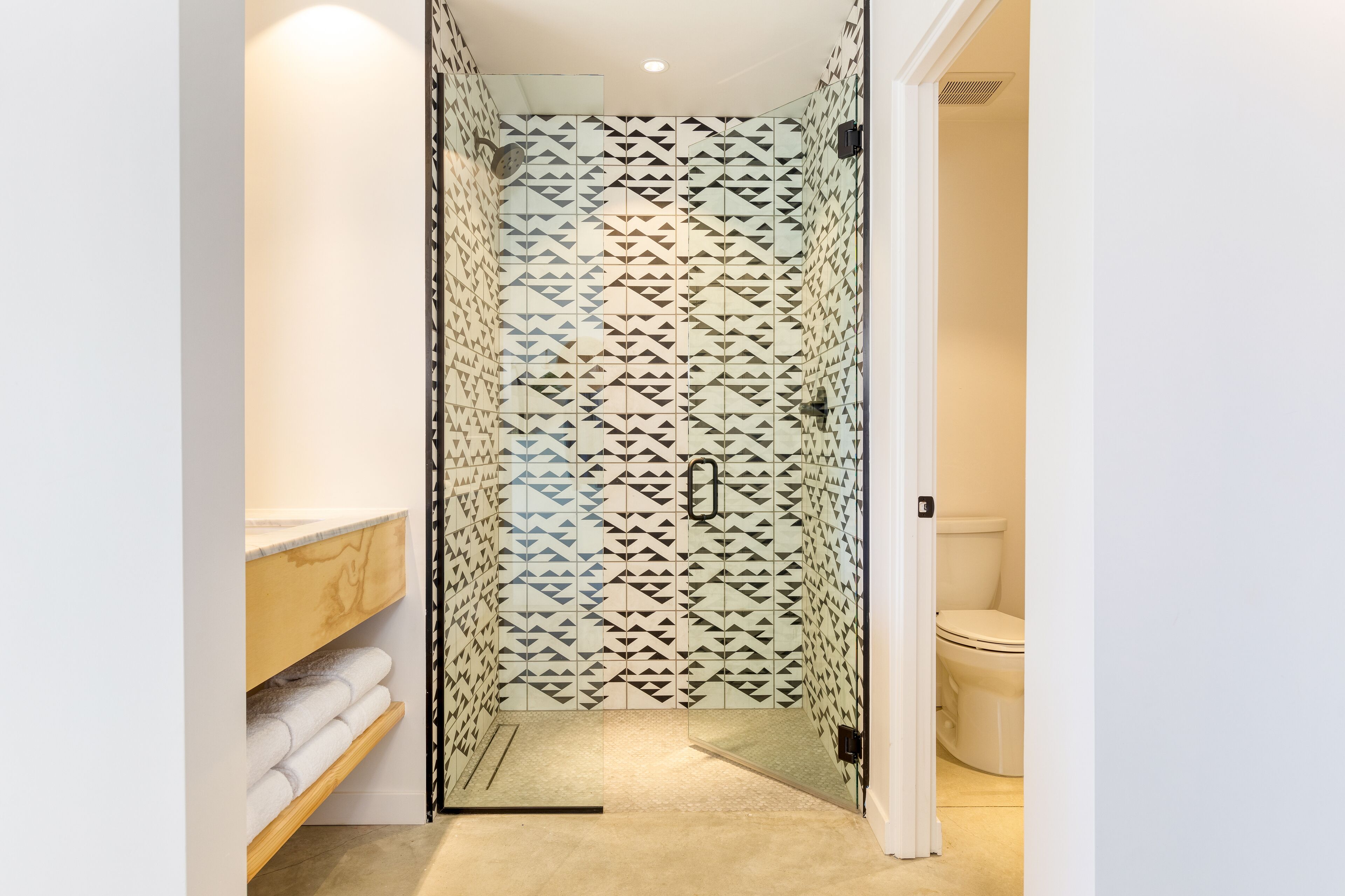 A walk-in shower with geometric patterned glass doors, a wooden shelf with towels, and a white toilet visible through the door.