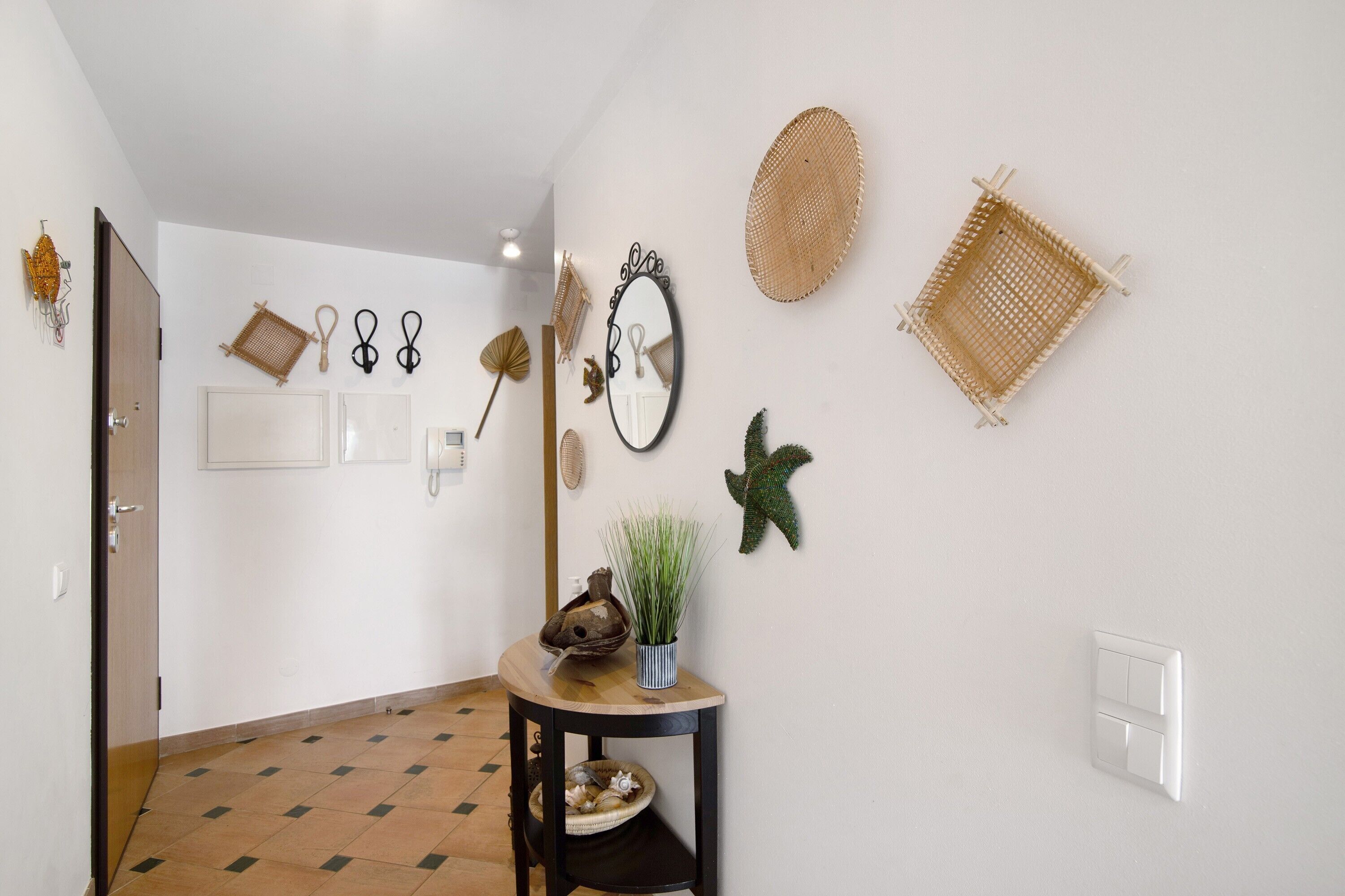 A hallway with a tiled floor, a door, a small table with a plant, and various wall decorations including a mirror, starfish, and woven baskets.