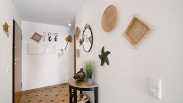 A hallway with a tiled floor, a door, a small table with a plant, and various wall decorations including a mirror, starfish, and woven baskets.