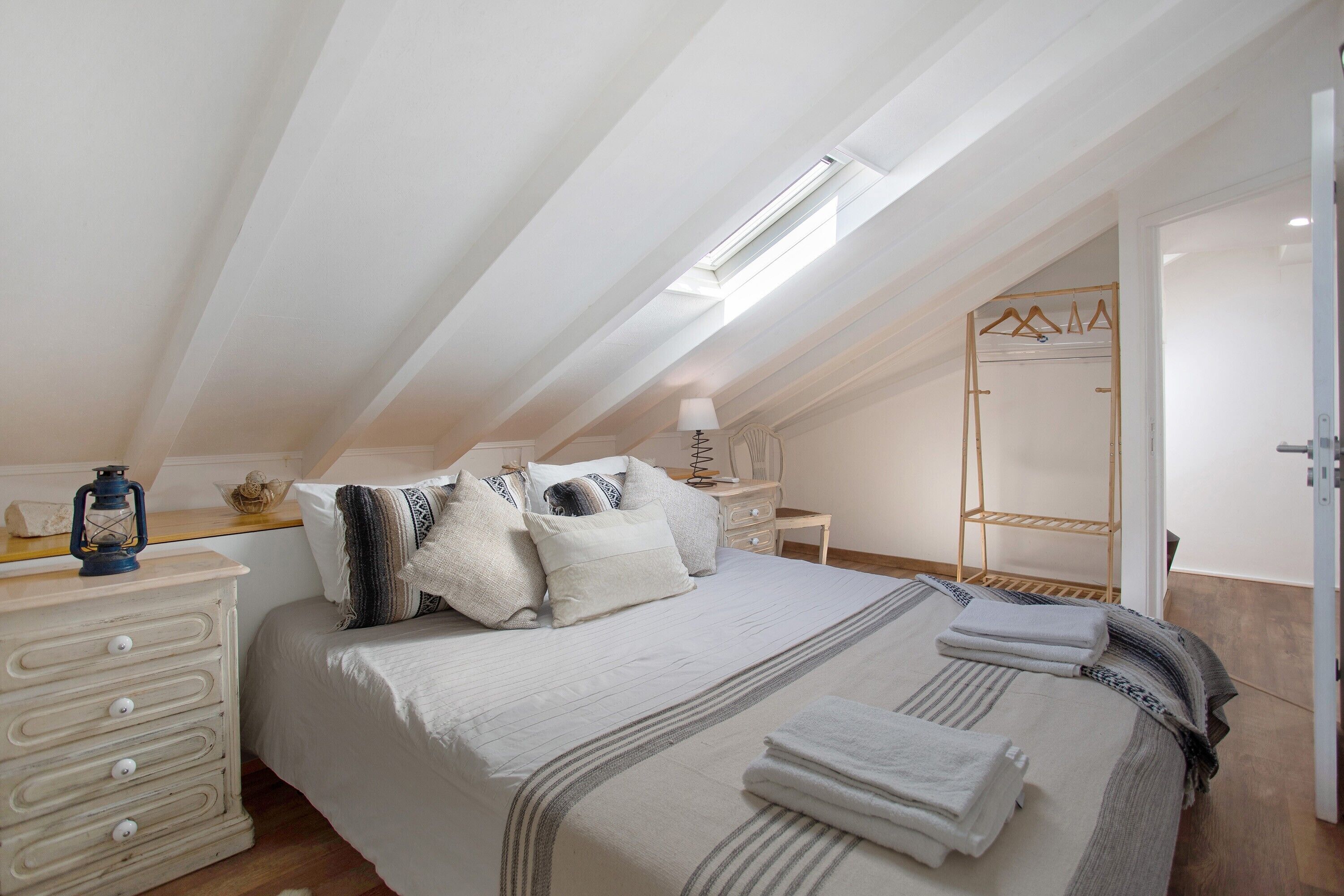 Ein Schlafzimmer mit schrägem Dach, einem Bett mit Kissen, einem Nachttisch mit Lampe, einem Holzregal und einem Kleiderständer.