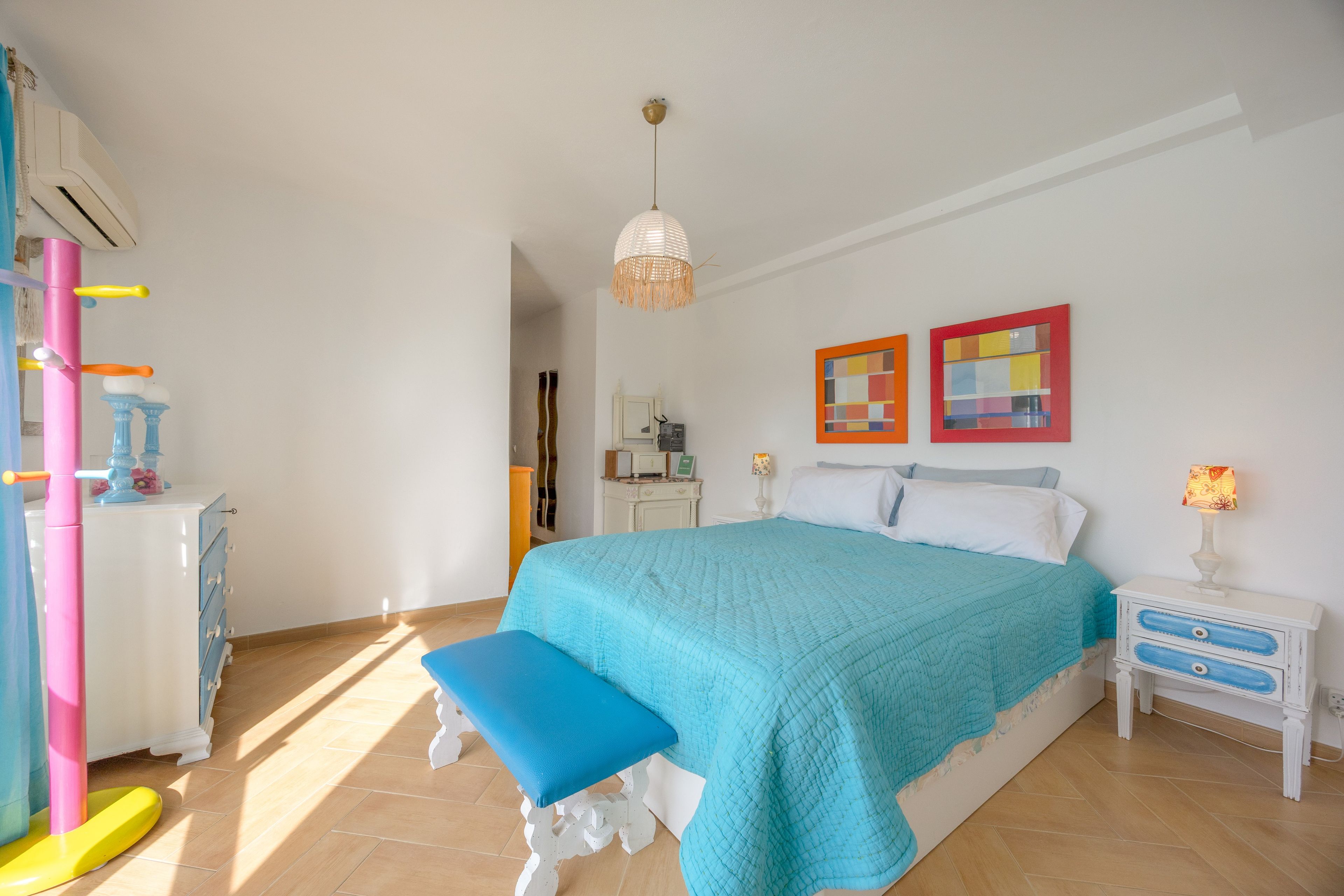 Quarto com cama, um banquinho, mesa de cabeceira, luminária e ar-condicionado fixado na parede.
