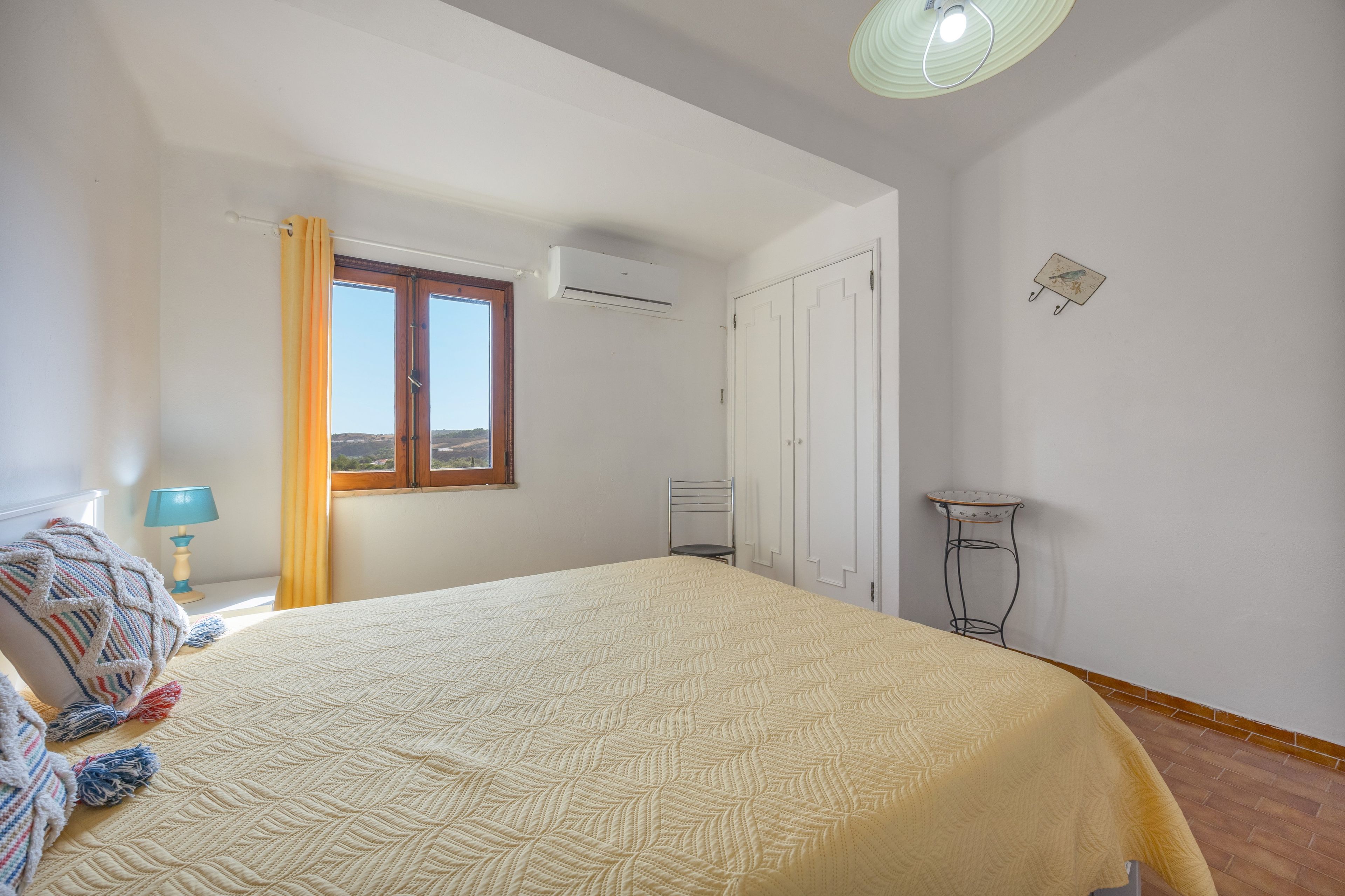 Une chambre à coucher comprenant un lit, une fenêtre, un appareil de climatisation, une table de chevet avec une lampe et une étagère fixée au mur.