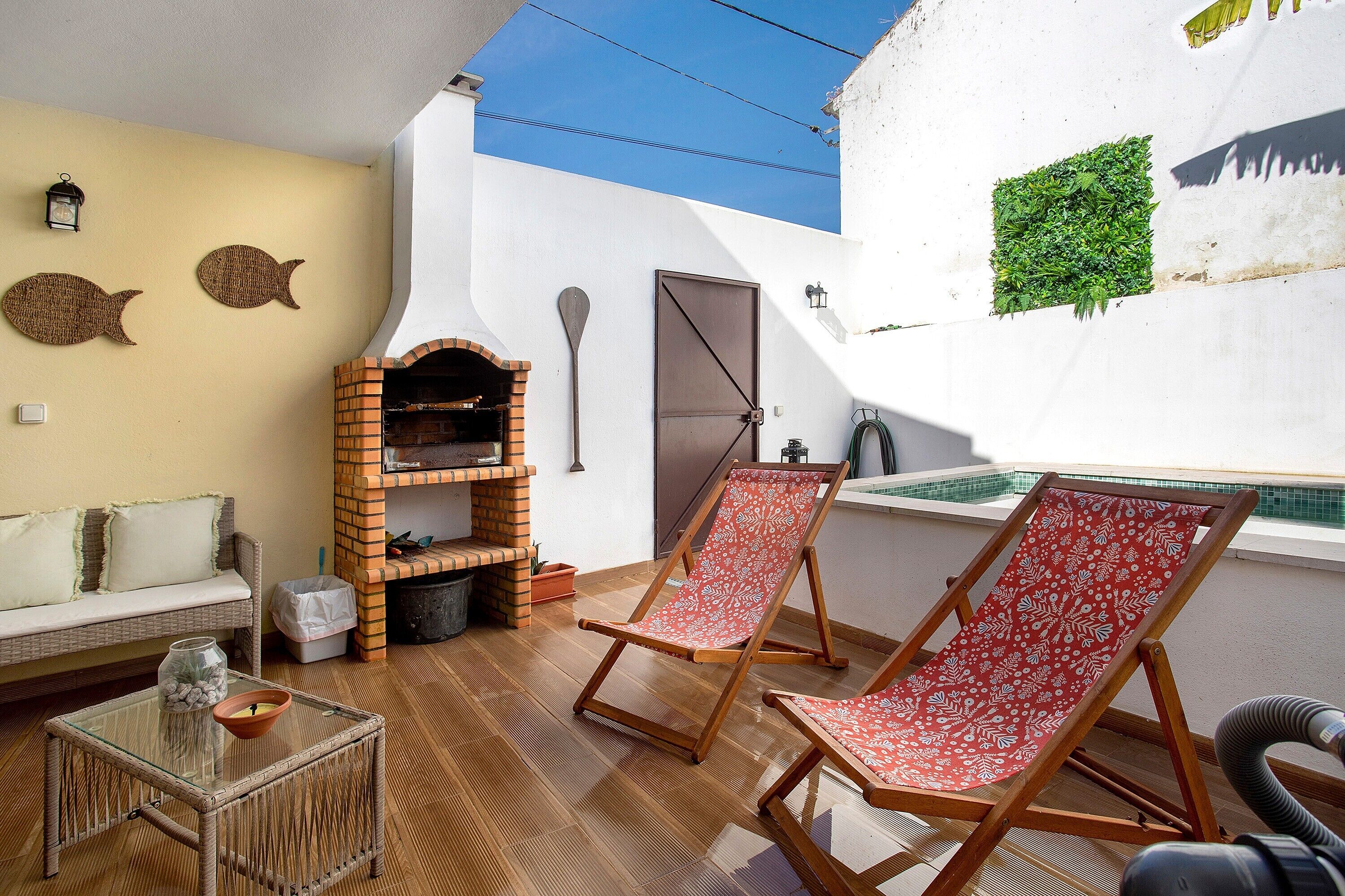 A patio with a brick oven, two red and white patterned lounge chairs, a glass table with a jar and bowl, a white wall with fish decorations, and a small green plant.