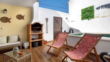 A patio with a brick oven, two red and white patterned lounge chairs, a glass table with a jar and bowl, a white wall with fish decorations, and a small green plant.