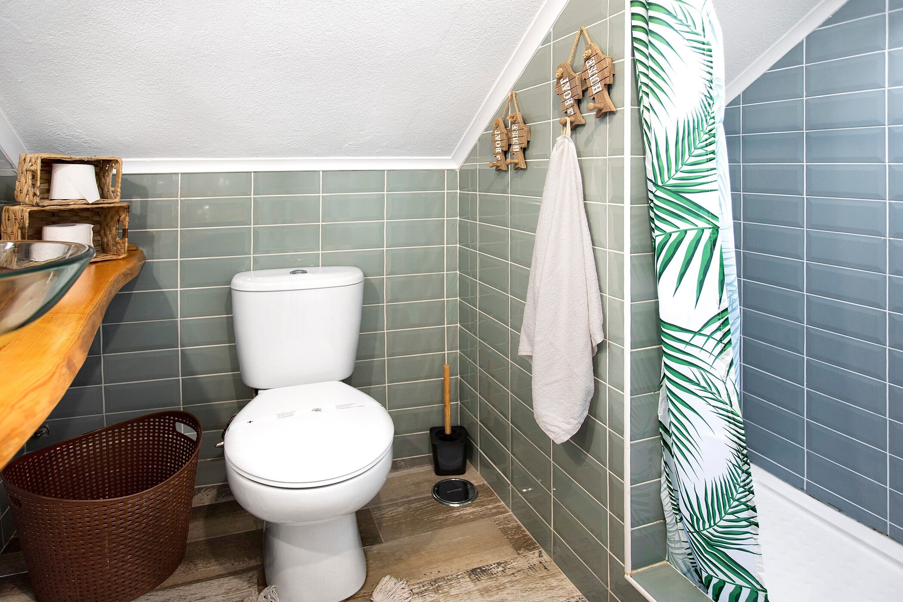 Ein Badezimmer mit Toilette, einem Korbwäscher, einer Holzablage mit Toilettenartikeln und einer Duschvorhang mit Blättermuster.