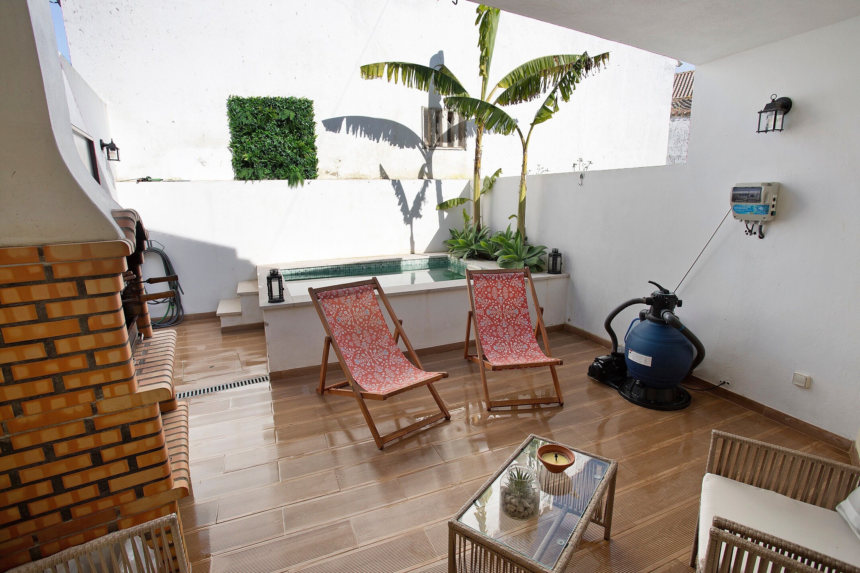 A room with a wooden floor, two red patterned chairs, a glass table with a candle, a blue vacuum cleaner, and a small window with a view of a white wall and a green plant.