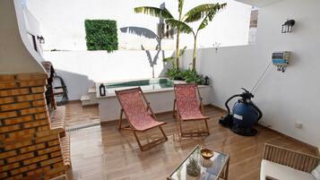 A room with a wooden floor, two red patterned chairs, a glass table with a candle, a blue vacuum cleaner, and a small window with a view of a white wall and a green plant.