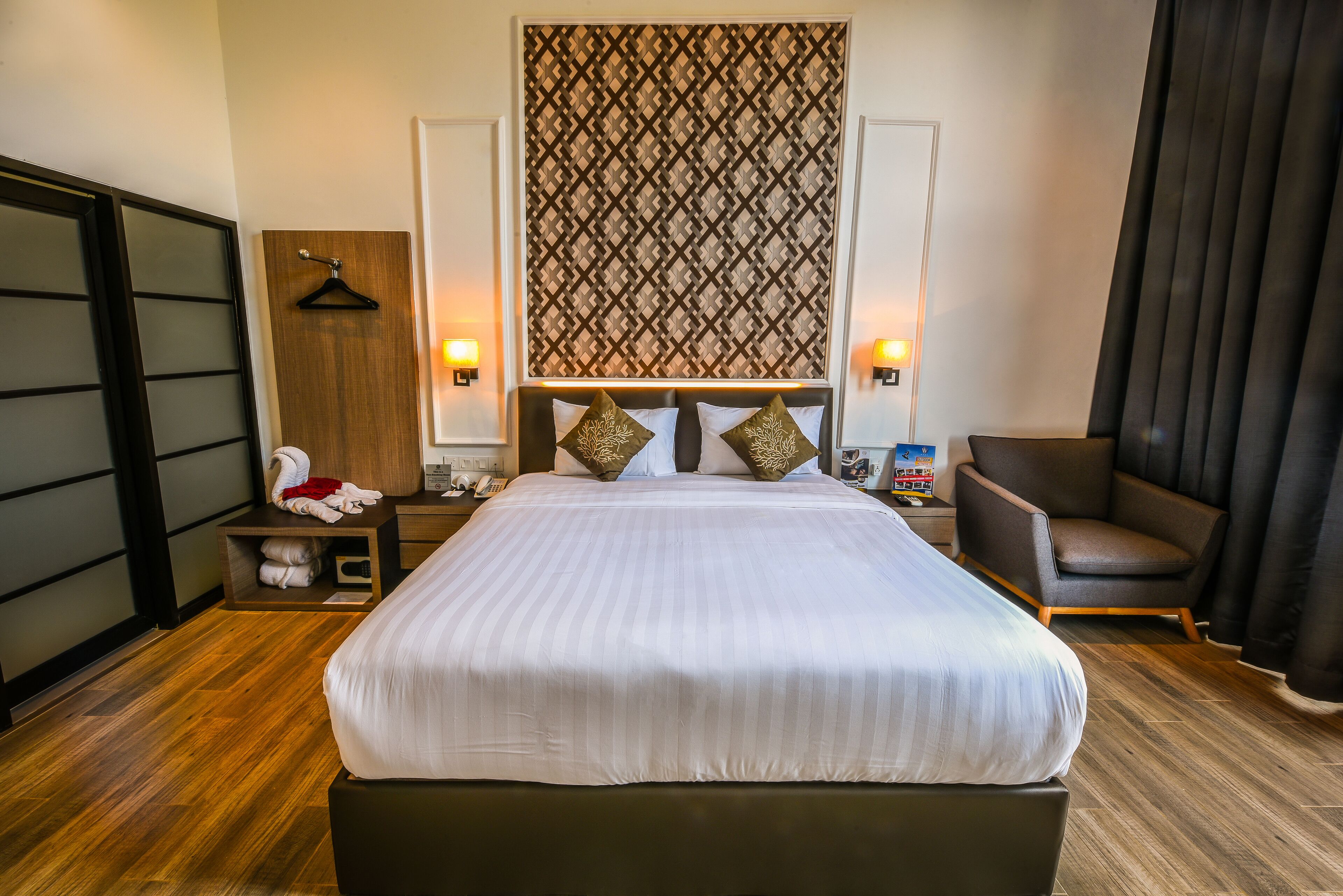 A hotel room with a large bed, a wooden headboard, a hanging coat rack, a side table with a lamp, and a chair.