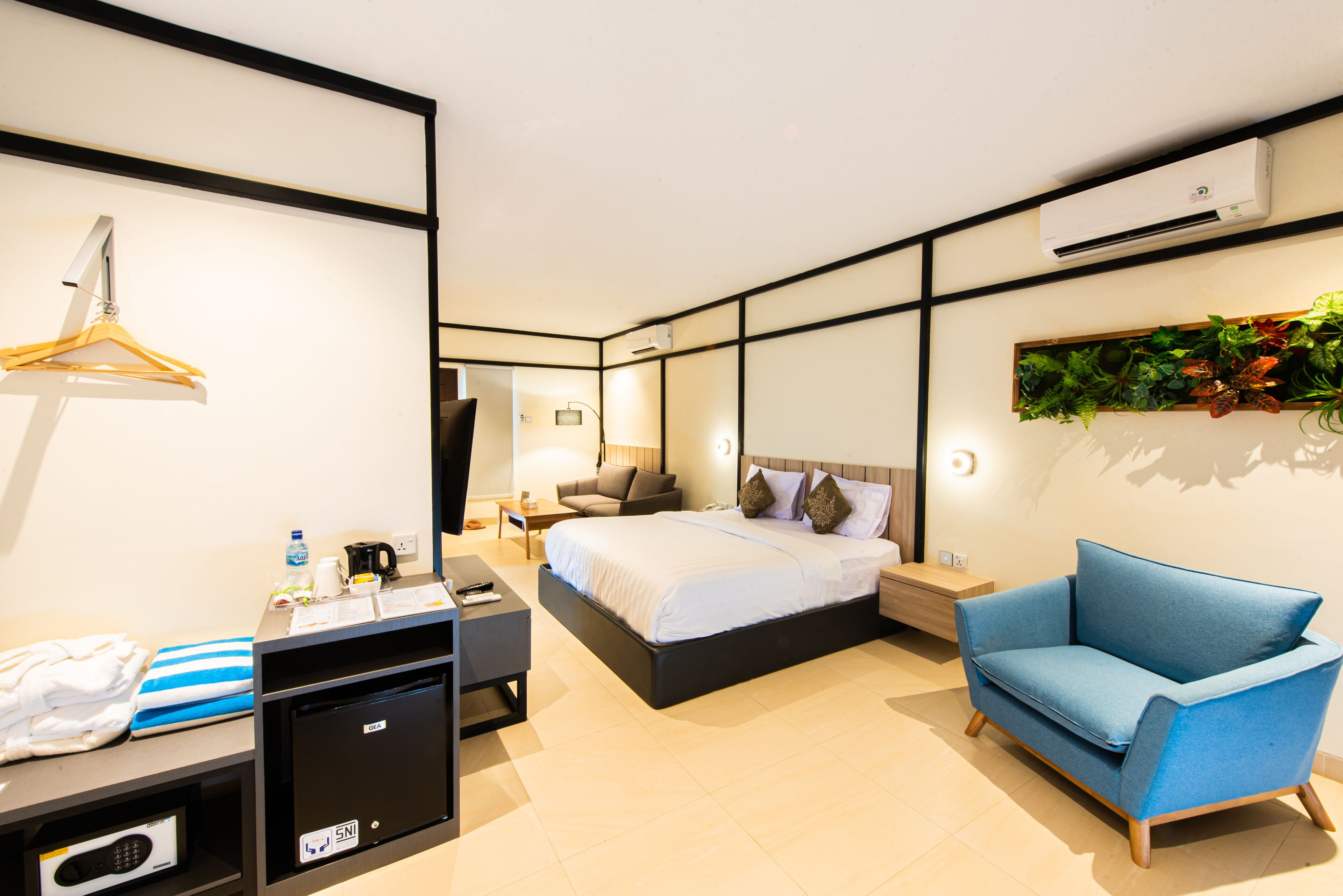 A hotel room with a bed, a blue armchair, a desk with a computer, a mini fridge, and a hanging coat rack.