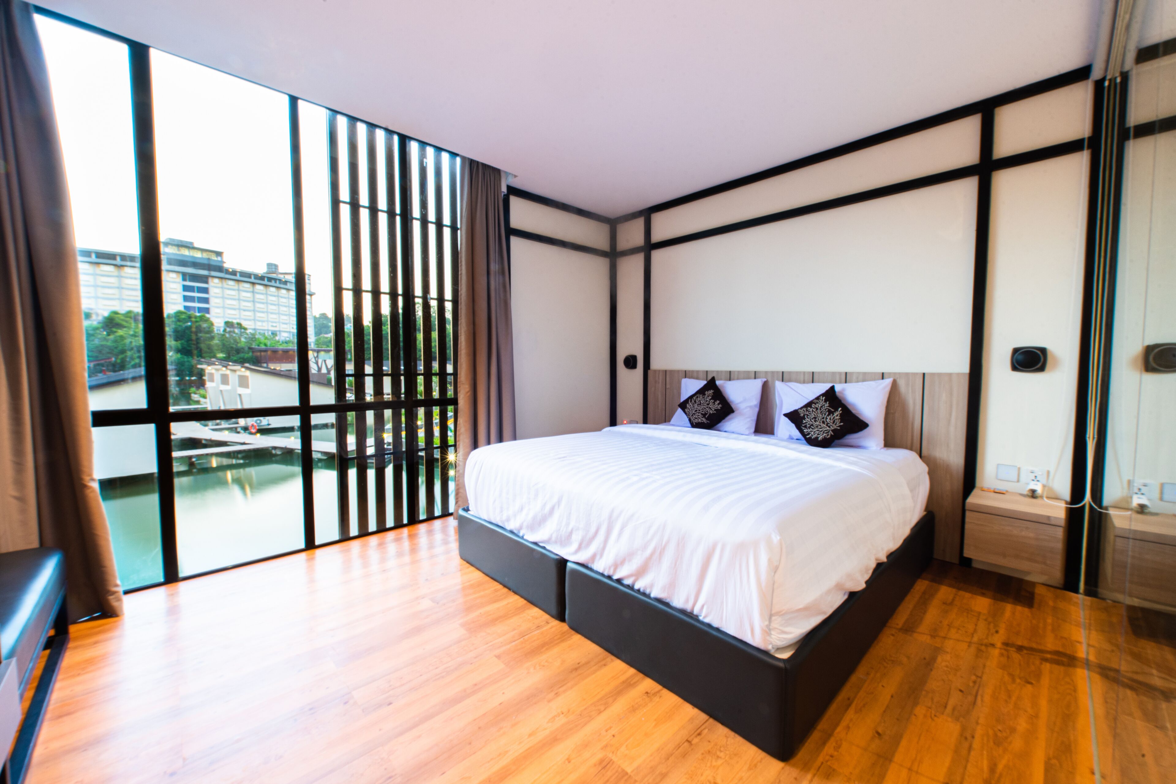 A modern bedroom with a large bed, wooden flooring, and a view of a cityscape through floor-to-ceiling windows.