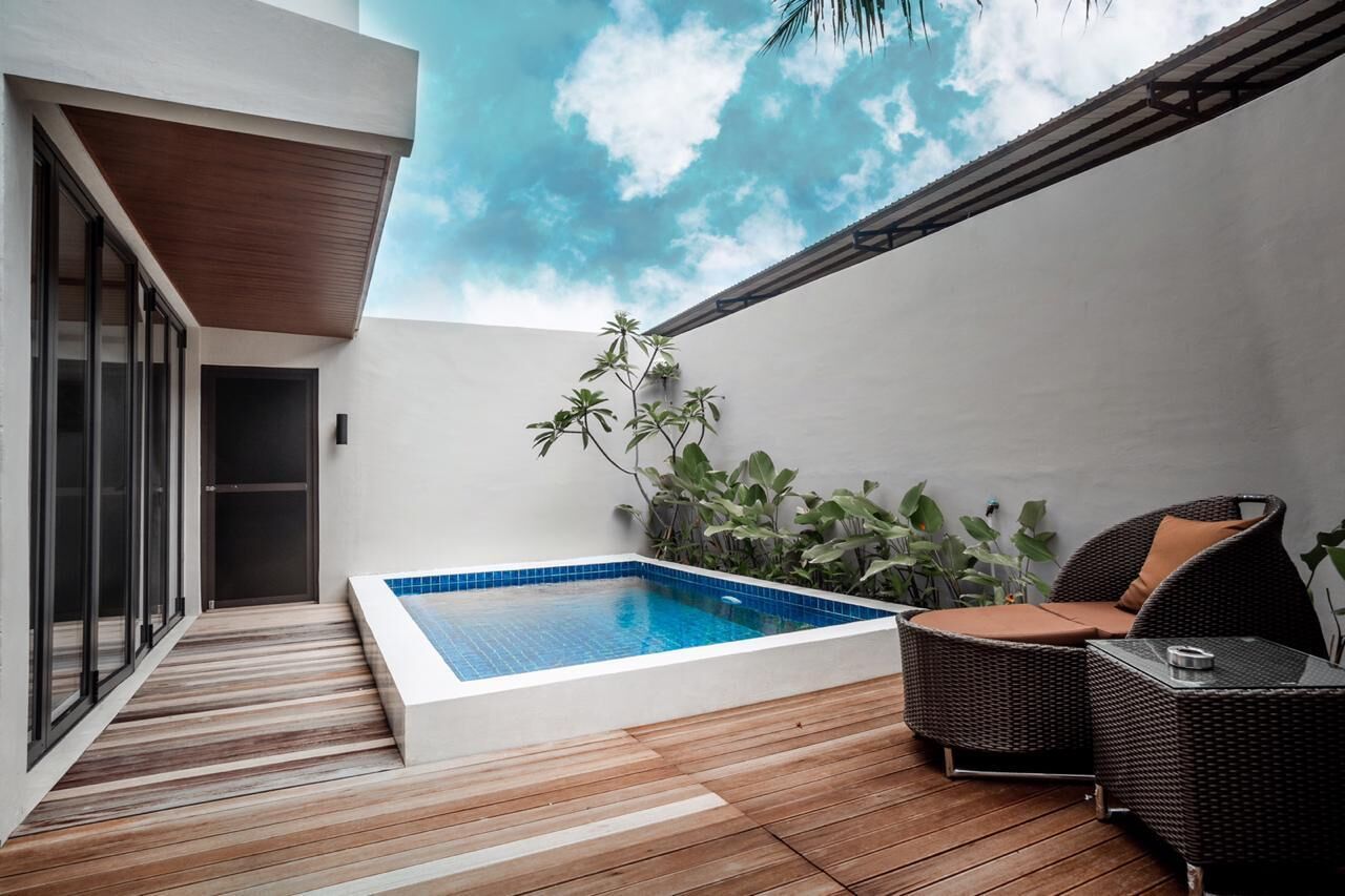 A modern outdoor pool area with a wooden deck, a small seating area with wicker furniture, and a view of the sky through a ceiling opening.