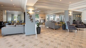 A spacious living area with a grey sectional sofa, a grey armchair, and a grey sofa. There is a large potted plant and a small table with a vase of flowers. The room has a tiled floor and a high ceiling with recessed lighting. - Flegra Palace Hotel & Spa (Kassandra)