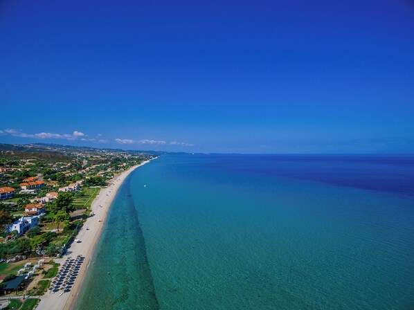 An aerial view of a coastal area with a long sandy beach, clear blue waters, and a mix of residential buildings along the shore. - Flegra Palace Hotel & Spa (Kassandra)