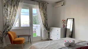 Un dormitorio con cama, una silla, una mesita, un aparador y una ventana con cortinas.
