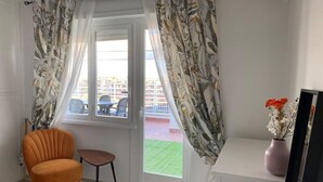 Una habitación con ventana, una silla, una mesita y un florero con flores.