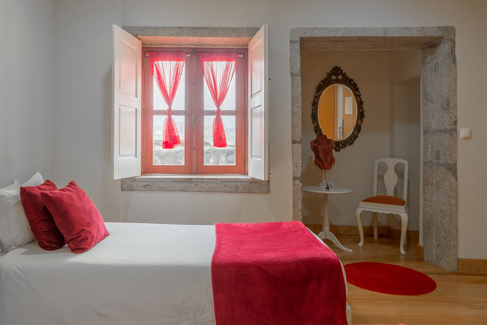 A bedroom with a bed, a window with red curtains, a chair, and a mirror.