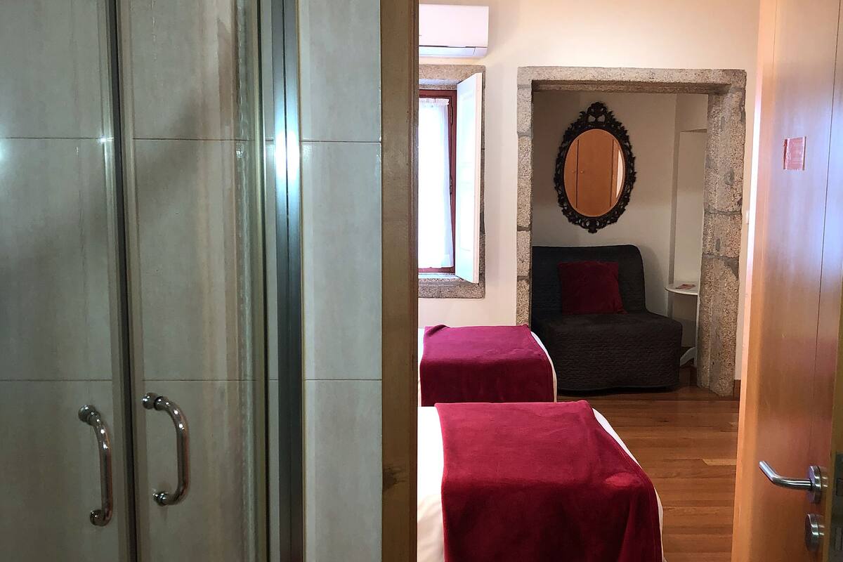 A hotel room with a glass shower enclosure, two beds with red bedding, a wooden floor, and a stone wall with a mirror.