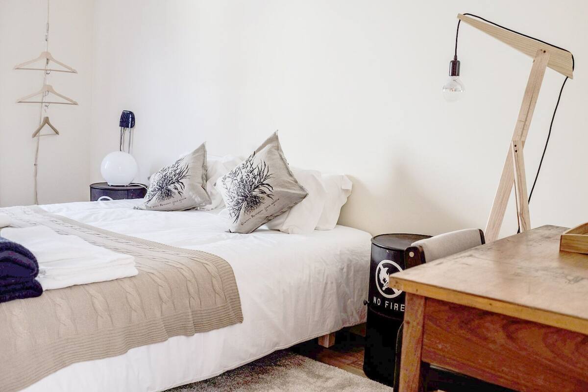 A neatly made bed with a white comforter and two decorative pillows, a bedside table with a lamp, and a small black trash bin with a fire safety symbol.