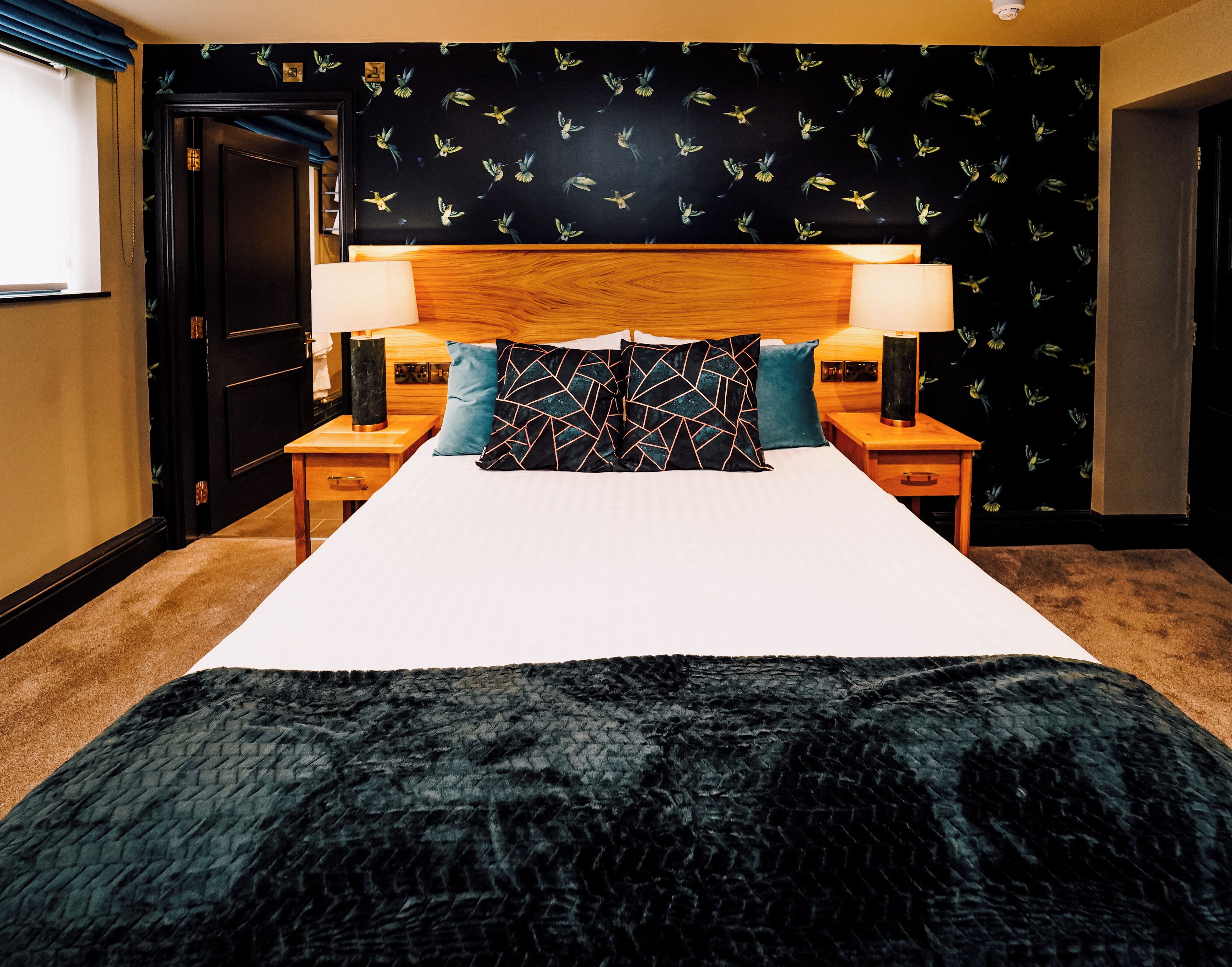 A hotel room with a large bed, two bedside tables, a wooden headboard, and a black wall with a bird pattern.
