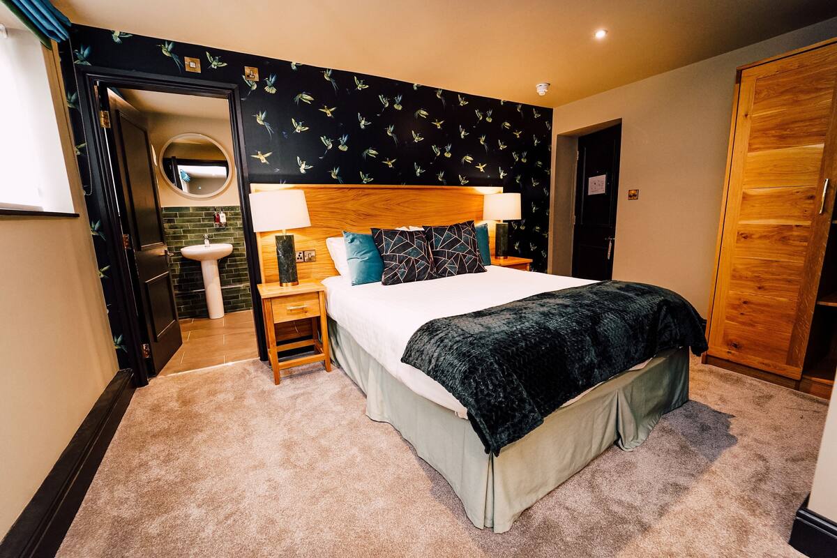 A hotel room with a bed, bedside lamps, a round mirror, a wooden wardrobe, and a bathroom with a sink.