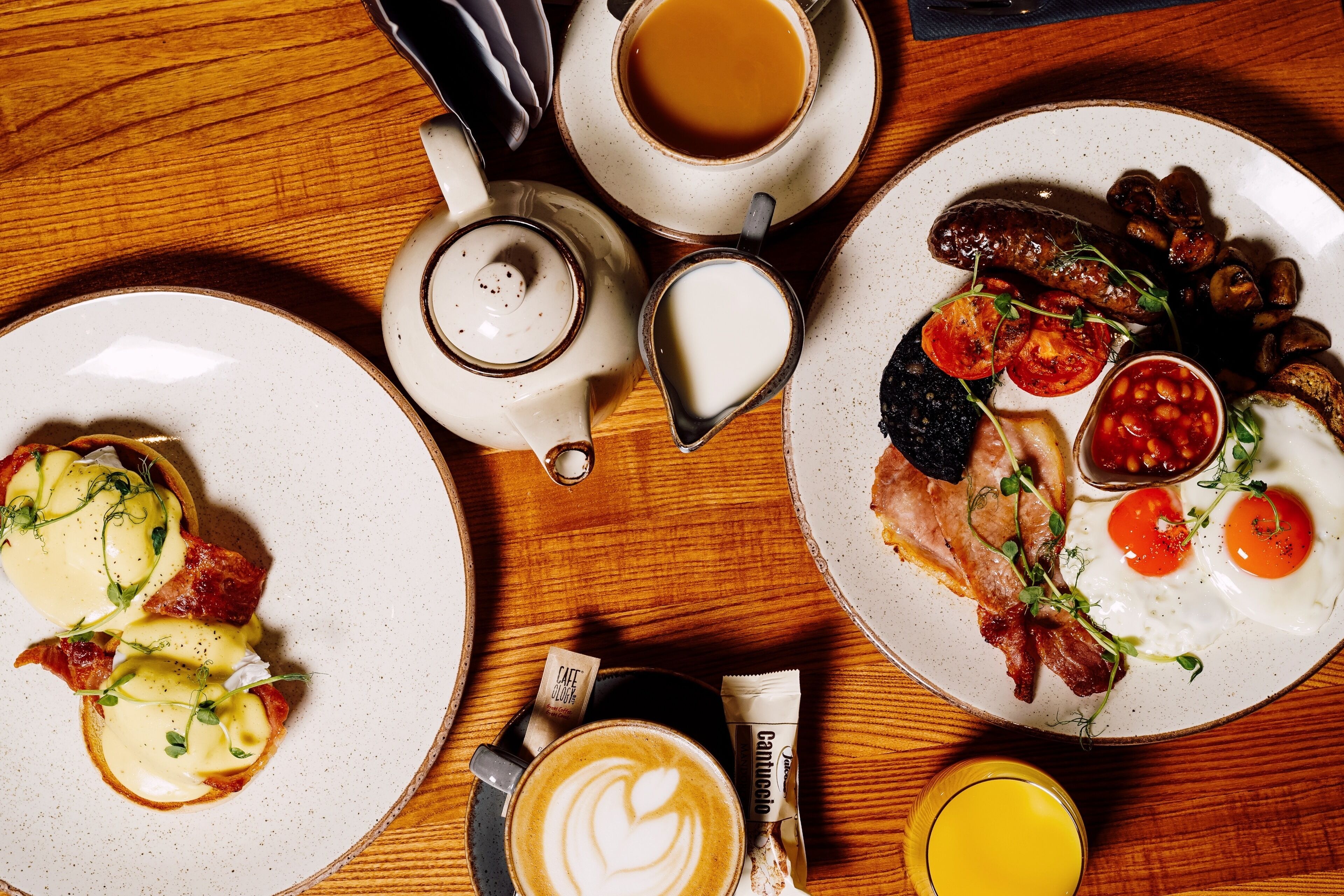 A breakfast setting with plates of eggs, bacon, sausages, and toast, a teapot, a cup of coffee, and a glass of orange juice.
