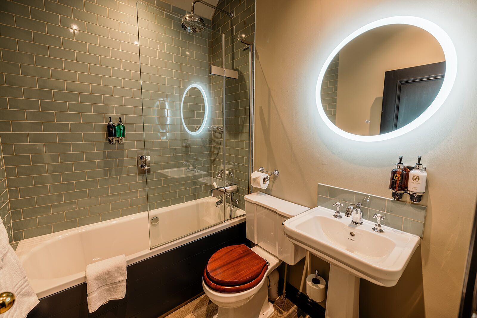 Un bagno con doccia vetrata, vasca, WC con sedile in legno, lavandino e specchio con illuminazione circolare.