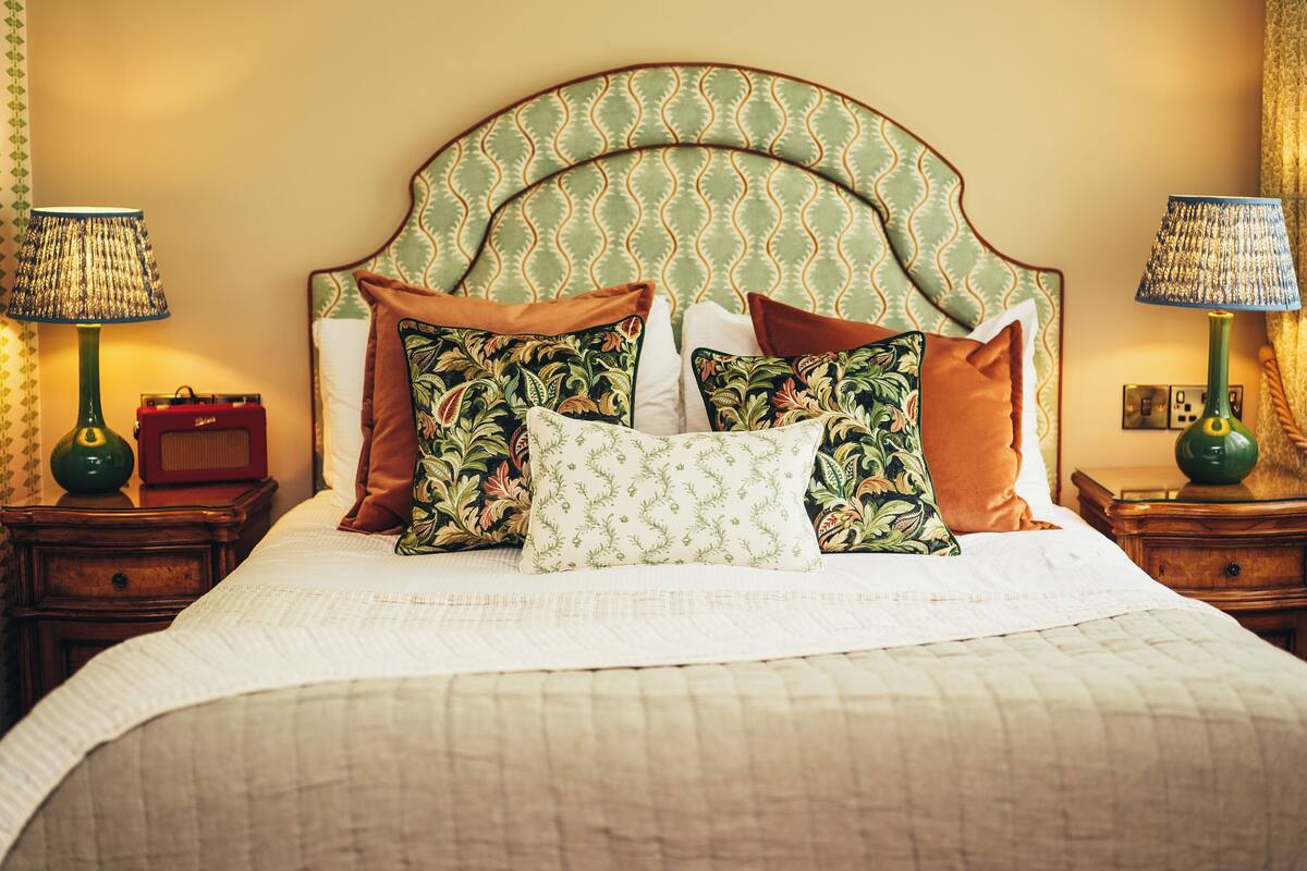 A neatly made bed with patterned pillows and a green headboard. There are two bedside tables with lamps and a small clock on one.