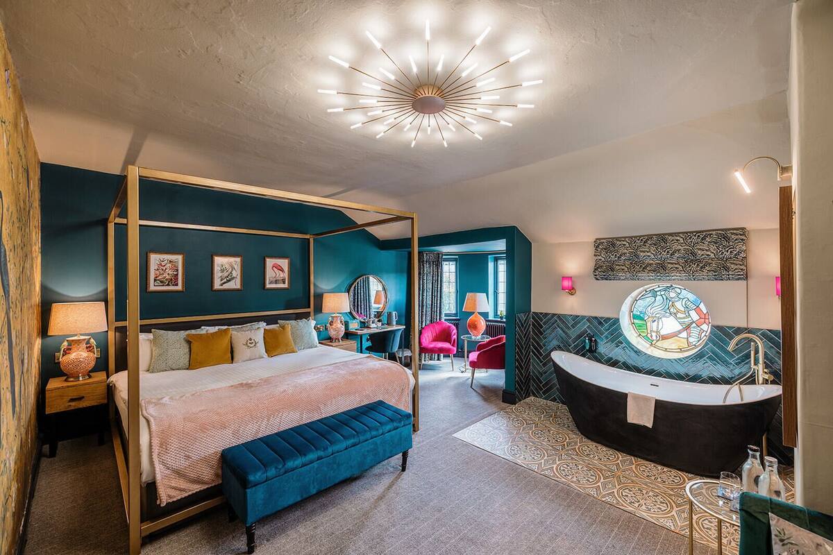 A modern bedroom with a four-poster bed, a large window, a sitting area with a chair and table, and a bathroom with a freestanding bathtub.