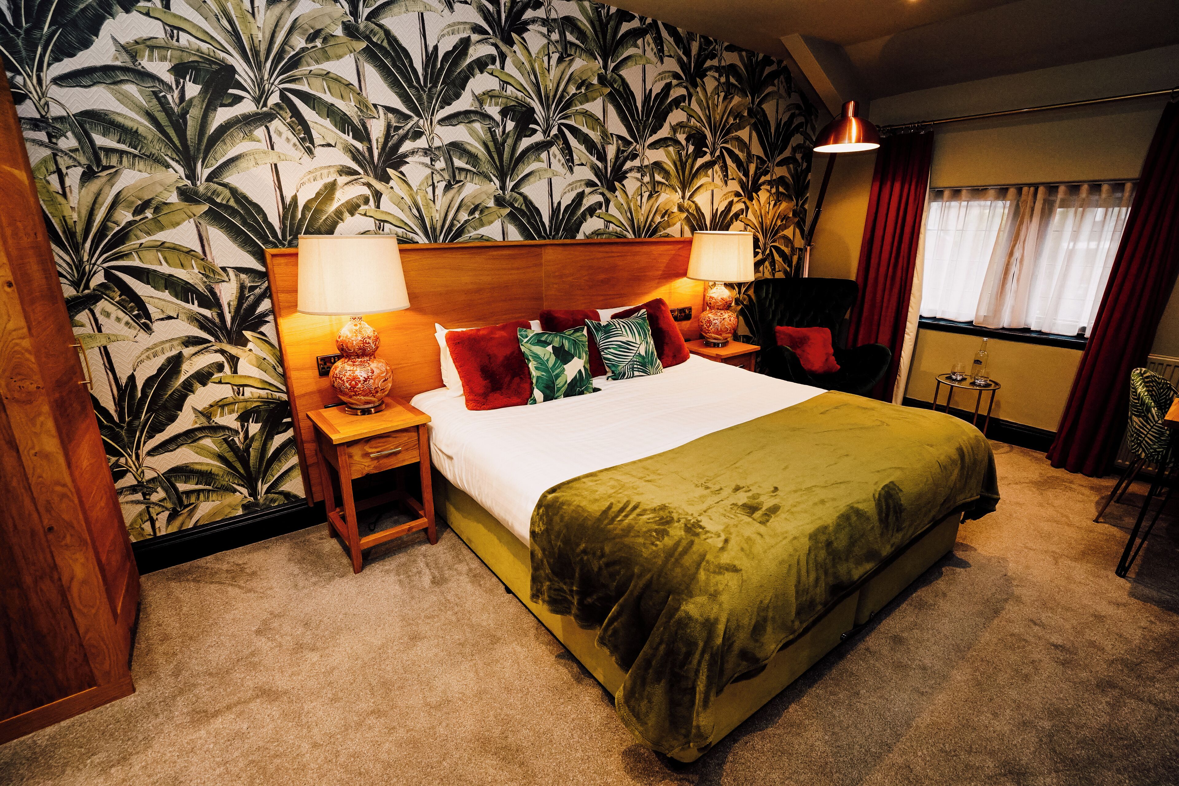 Una habitación de hotel con una cama grande, dos mesitas de noche con lámparas, una pequeña mesa con lámpara, una silla y una ventana con cortinas.